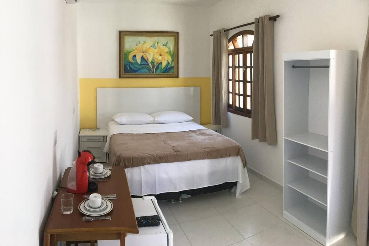 Ein Hotelzimmer mit einem Bett, einem Schreibtisch mit rotem Wasserkocher, einer weißen Klimaanlage, einem Deckenventilator und einem Fenster mit Vorhängen.