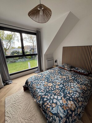 Ein Schlafzimmer mit großem Fenster, einem Bett mit Blumenmuster auf der Bettwäsche, einer Hängelampe und einer geneigten Decke.
