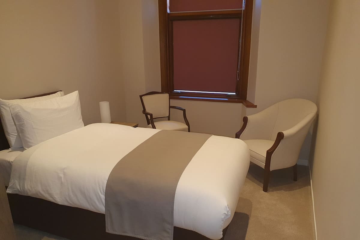 A bedroom with a bed, two chairs, a window, and a wall-mounted shelf.