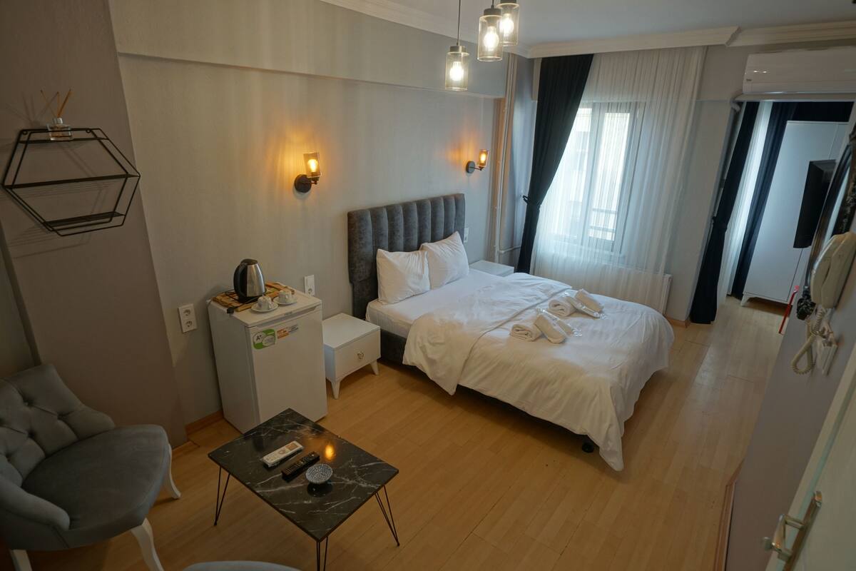 A hotel room with a bed, bedside table, chair, and a small desk with a kettle.