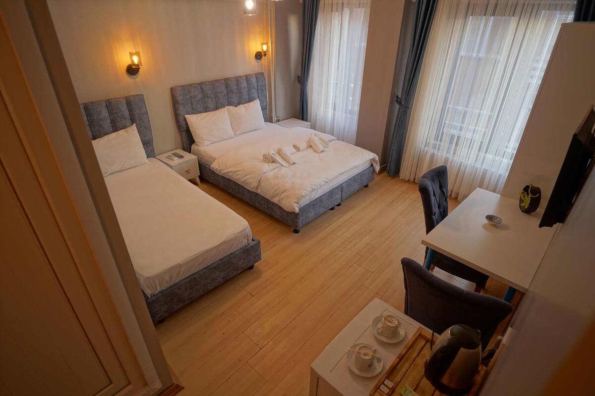 A hotel room with two beds, a desk, and a chair. There are two bedside tables with lamps and a small table with a kettle and cups.
