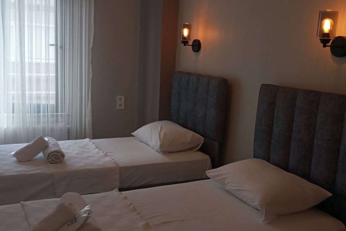 A hotel room with two beds, each with a pillow and a rolled towel. There are wall-mounted lights and a window with sheer curtains.