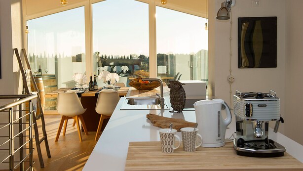 Ett modernt kök med ett stort fönster, en kaffemaskin och en matplats med stolar och ett bord som är dukat för två personer.