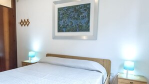 Un lit bien fait, recouvert d’une couette blanche, avec deux lampes de chevet, une œuvre d’art encadrée au mur et une table de chevet en bois dotée d’un tiroir.
