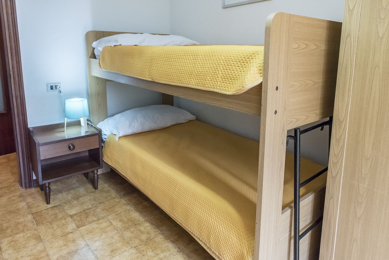 Un lit superposé avec une couverture jaune, une table de chevet avec une lampe et une petite table en bois.