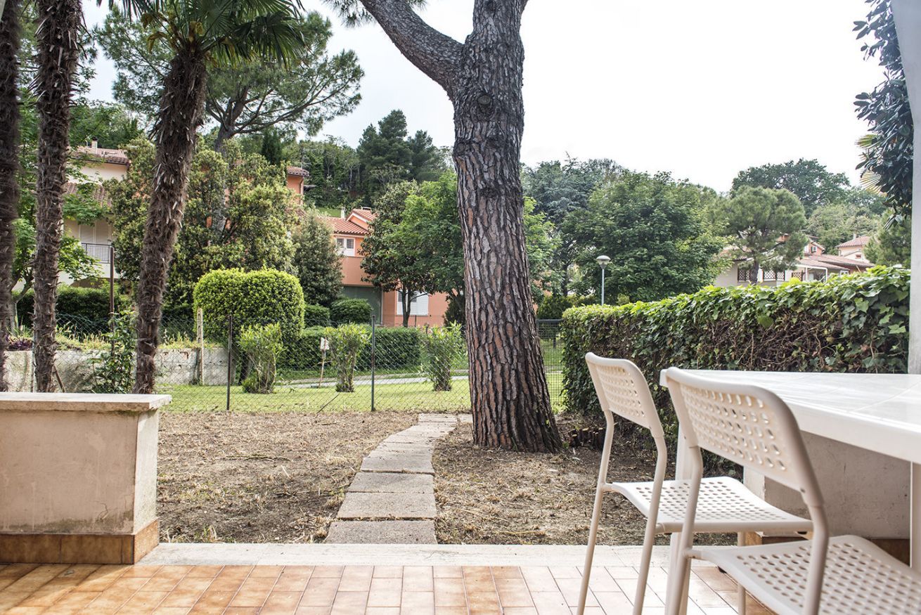 An outdoor patio area with a tiled floor, two white metal chairs, and a large tree.