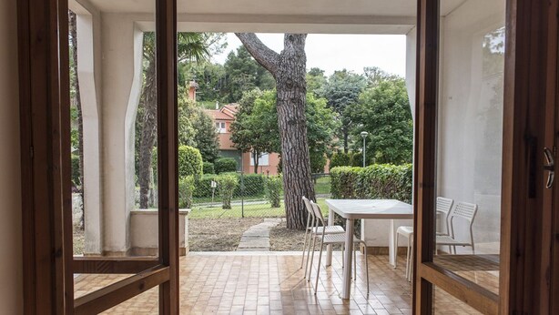 Une terrasse ouverte avec un sol carrelé, une table entourée de chaises et, en arrière-plan, une vue sur un arbre et des bâtiments.