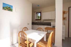 Un comedor con mesa blanca y sillas de madera, una cocina con armarios blancos, una ventana con vistas y un radiador.