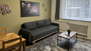 Een woonkamer met een donkergrijze bank, een houten tafel, een klein bijzettafeltje met een plant en een groot raam met gordijnen.