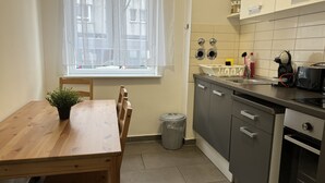 Een compacte keuken met een houten eettafel, een raam met dunne gordijnen, een afvalbak, en moderne apparaten waaronder een vaatwasser en een oven.