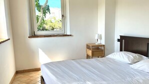 Un dormitorio con cama, mesita de noche, lámpara, ventana con vista a zonas verdes y piso de baldosas.