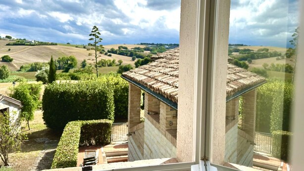 Vista desde una ventana sobre un paisaje rural con colinas onduladas, un edificio de piedra con tejado de tejas y un seto bien cuidado.
