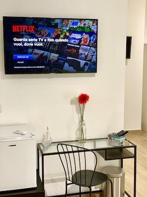 A living room with a flat-screen TV displaying a Netflix interface, a small table with a vase of flowers, a chair, and a trash can.
