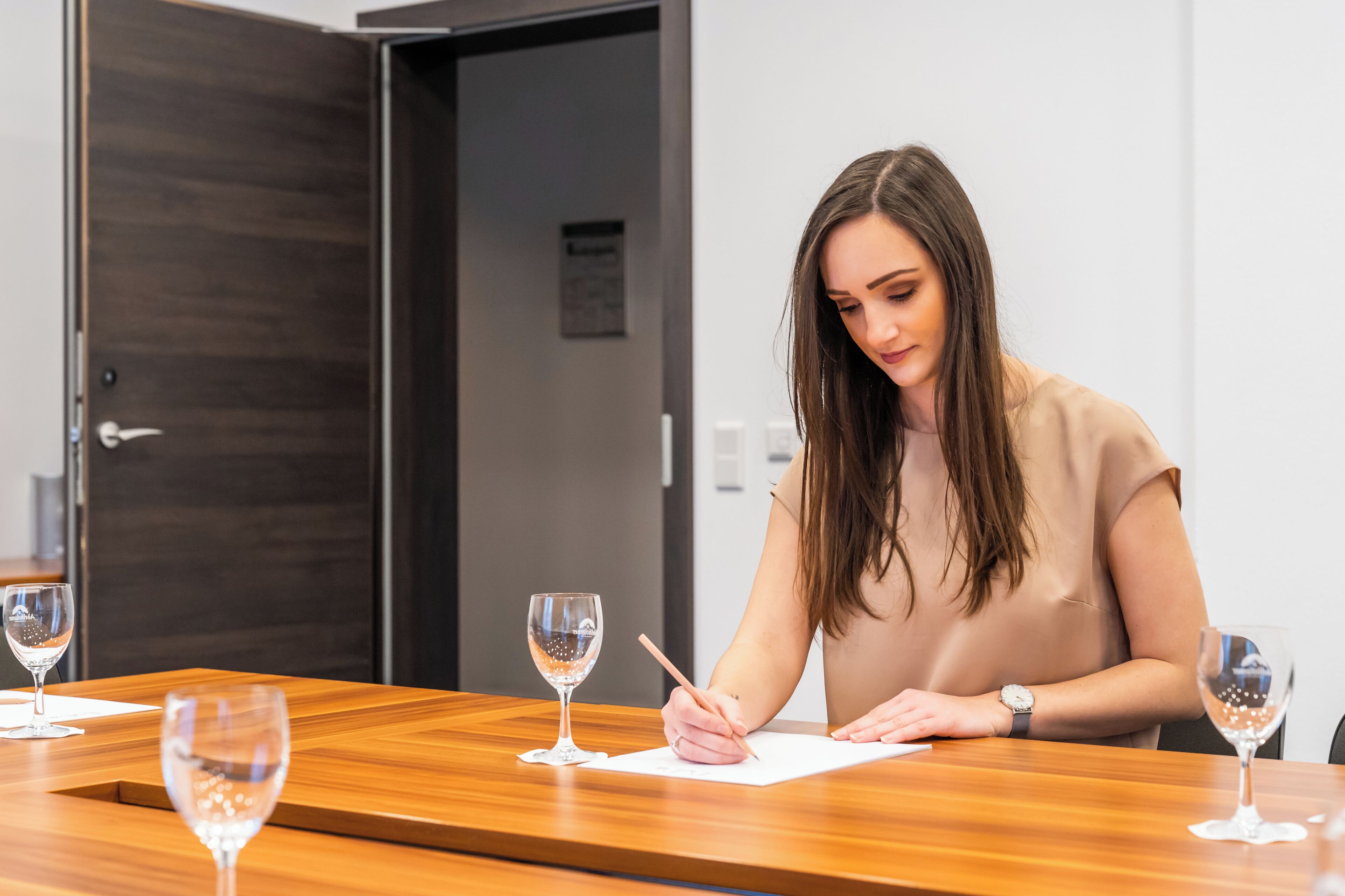 Una persona sentada a una mesa de madera con un bloc de notas y un bolígrafo, escribiendo. Hay tres copas de vino sobre la mesa.