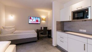 A compact hotel room with a bed, a desk, a chair, a television, and a kitchenette. - DuDa Boarding Apartments (Reichertshofen)