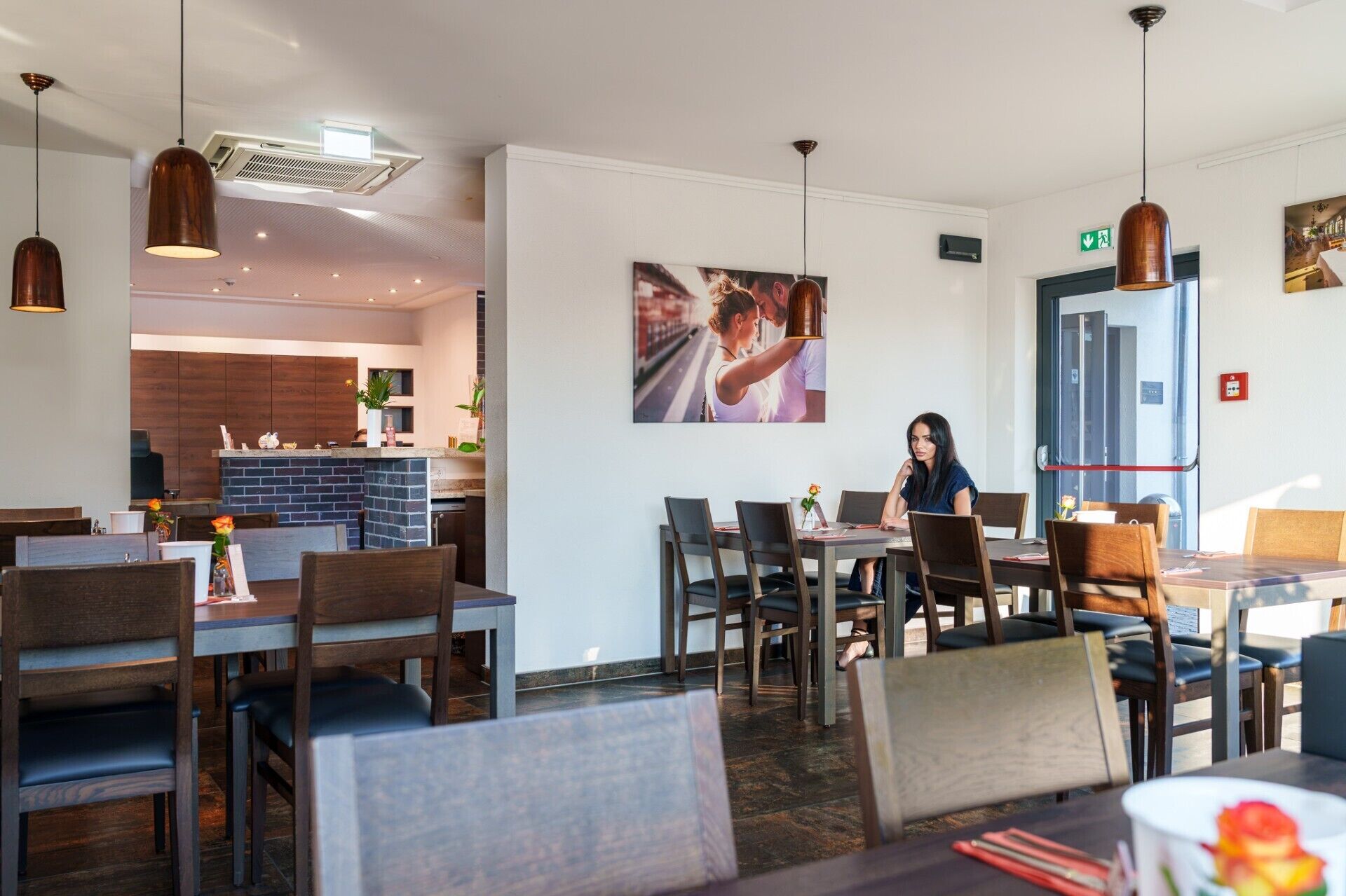 A restaurant interior with multiple dining tables and chairs, a kitchen area in the background, and a large framed picture on the wall.