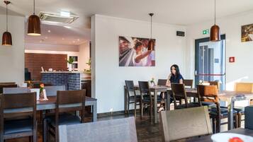 A restaurant interior with multiple dining tables and chairs, a kitchen area in the background, and a large framed picture on the wall.