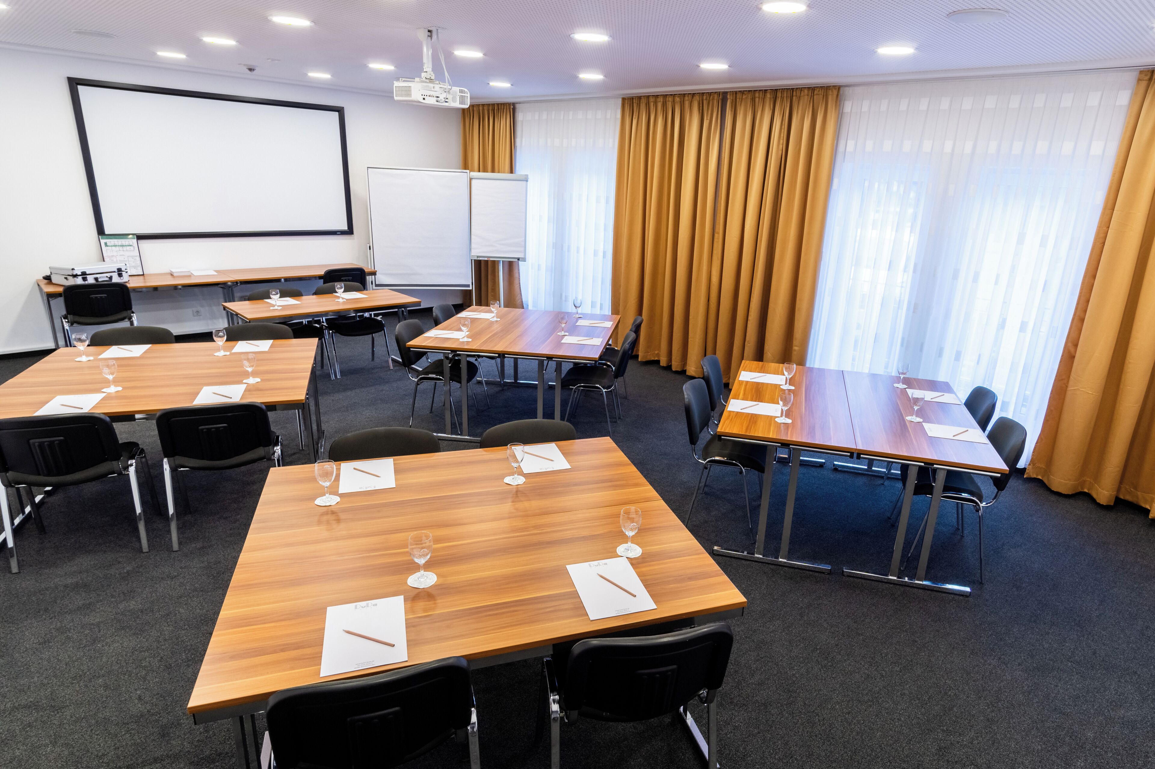 A conference room with multiple tables, chairs, a projector screen, and a large window with curtains.