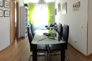 Un comedor con una mesa de vidrio y sillas negras, un sofá con cojines, una estantería y un ventanal con cortinas verdes.