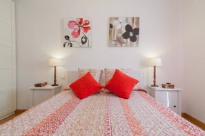 Una cama bien tendida con una colcha estampada y dos almohadas rojas, flanqueada por mesitas de noche con lámparas y una cajita en cada una.