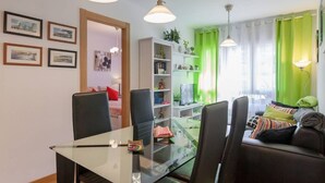 Ein modernes Wohnzimmer mit einem Glastisch, schwarzen Stühlen, einer Couch mit Kissen, einem Bücherregal und einem Fernseher.