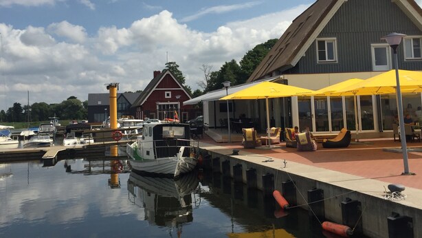 Un port de plaisance avec des bateaux amarrés, un bâtiment rouge avec un toit blanc et un espace de repos couvert agrémenté de parasols jaunes.