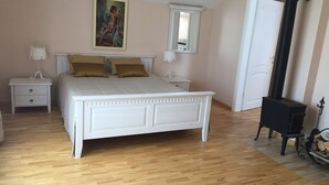 Une chambre avec un lit blanc, deux tables de chevet, une commode, un poêle noir et un tableau accroché au mur.