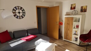 Un salón moderno con un sofá gris, una mesa de centro blanca y una silla roja. Hay un mueble de madera con libros y un reloj en la pared.