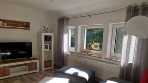 Un salón moderno con un televisor de pantalla plana, una estantería blanca, sofás grises y una ventana con vistas a zonas verdes.