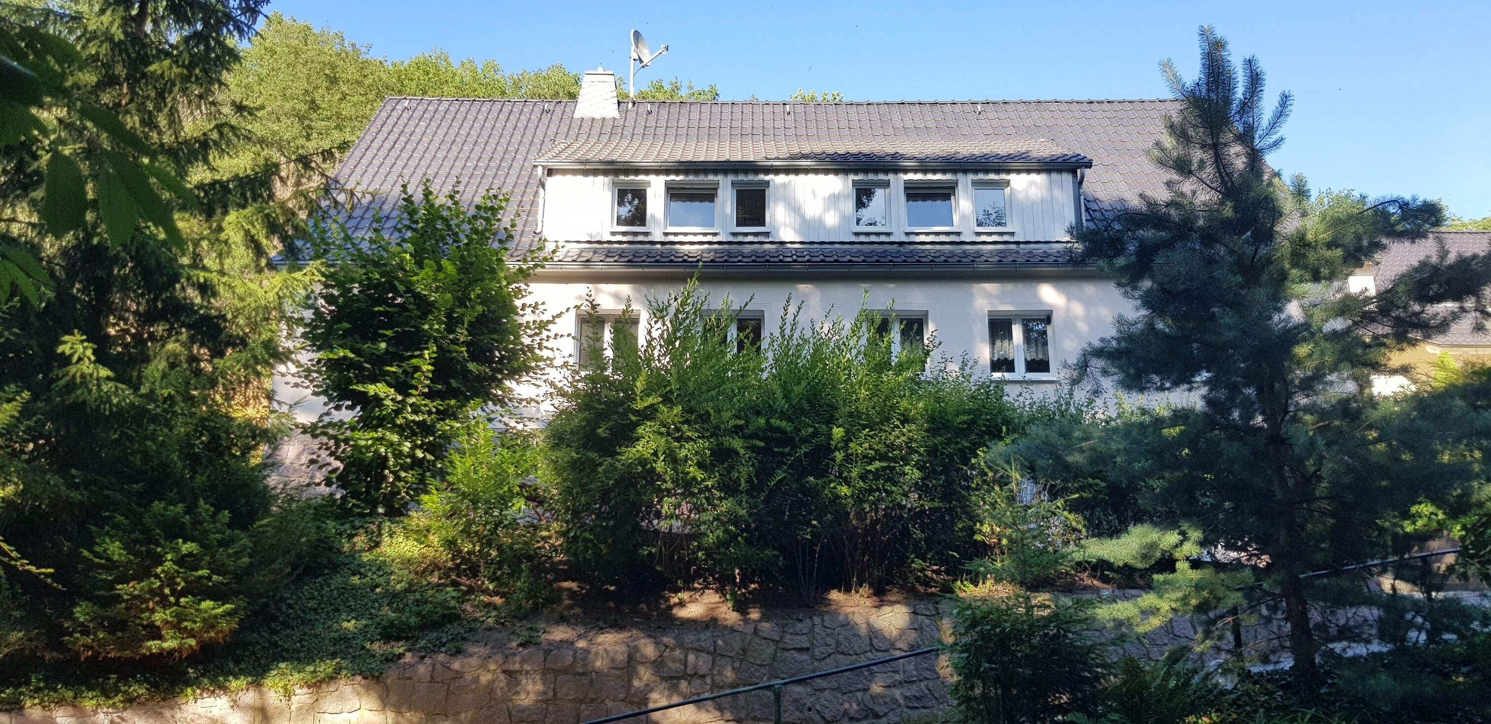 Una casa de dues plantes amb un mur de pedra al davant, envoltada de vegetació abundant.