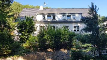 Una casa de dues plantes amb un mur de pedra al davant, envoltada de vegetació abundant.