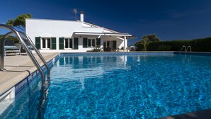 Un espace piscine moderne avec une piscine à l’eau d’un bleu limpide, un bâtiment blanc aux volets verts et une échelle en métal.