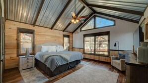 Ein Schlafzimmer mit Holzboden, einem Bett mit grauer Tagesdecke, einem Nachttisch, einem Schreibtisch mit Lampe und einem Deckenventilator.