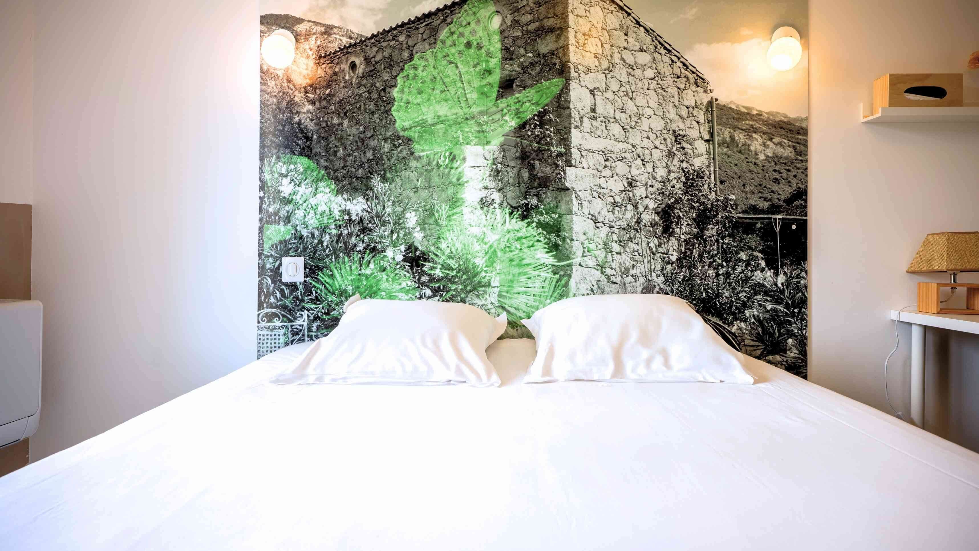 Un dormitorio con una cama grande, ropa de cama blanca y un mural en la pared que representa una pared de piedra y follaje.