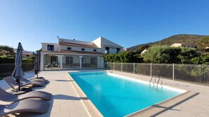 A swimming pool with lounge chairs and umbrellas, a fenced patio, and a building with a tiled roof. - Casa Nova Maison d'hotes (Coggia)