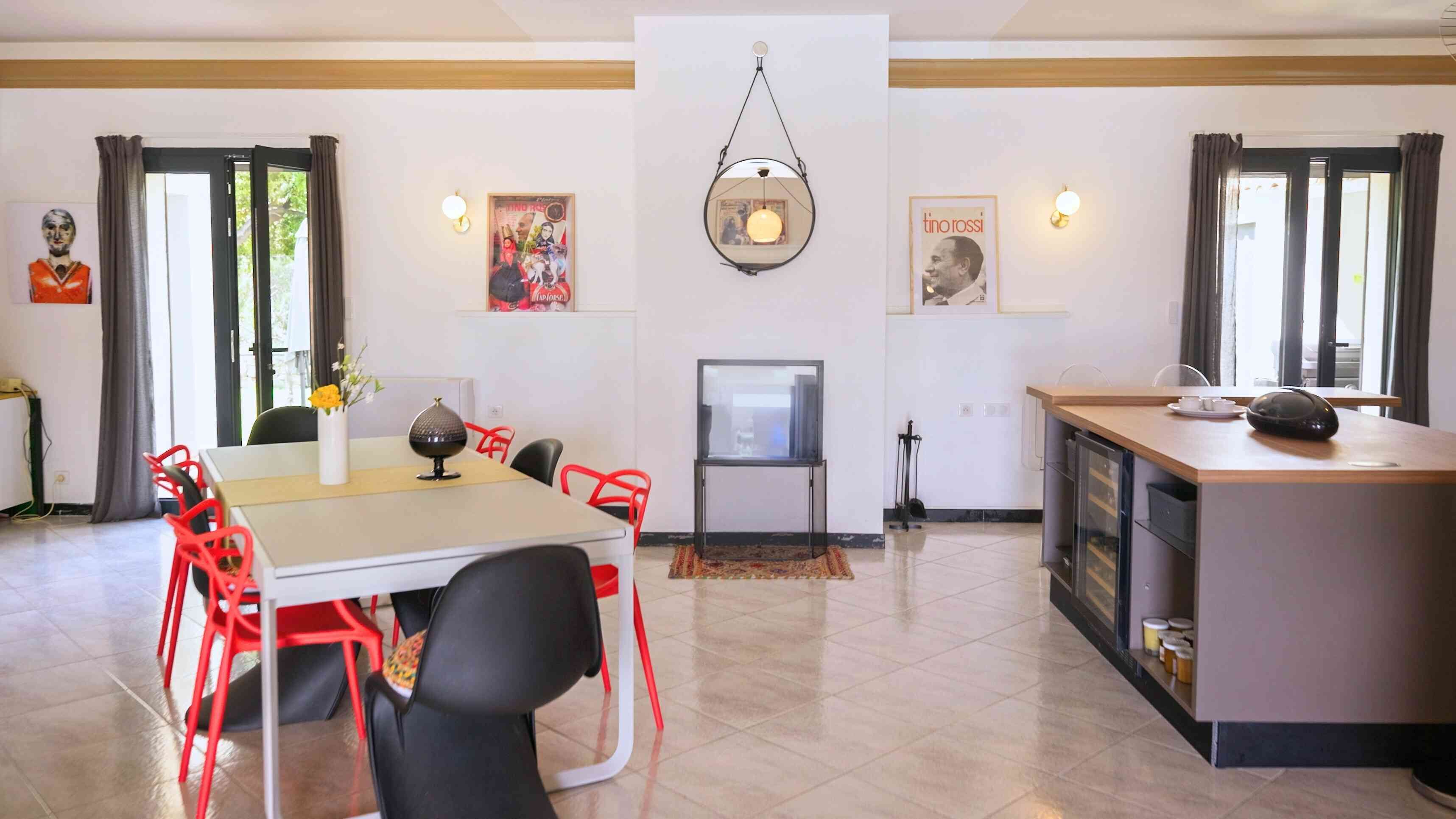 A modern kitchen with a dining area, featuring a central island, red chairs, a TV, and wall-mounted lights.