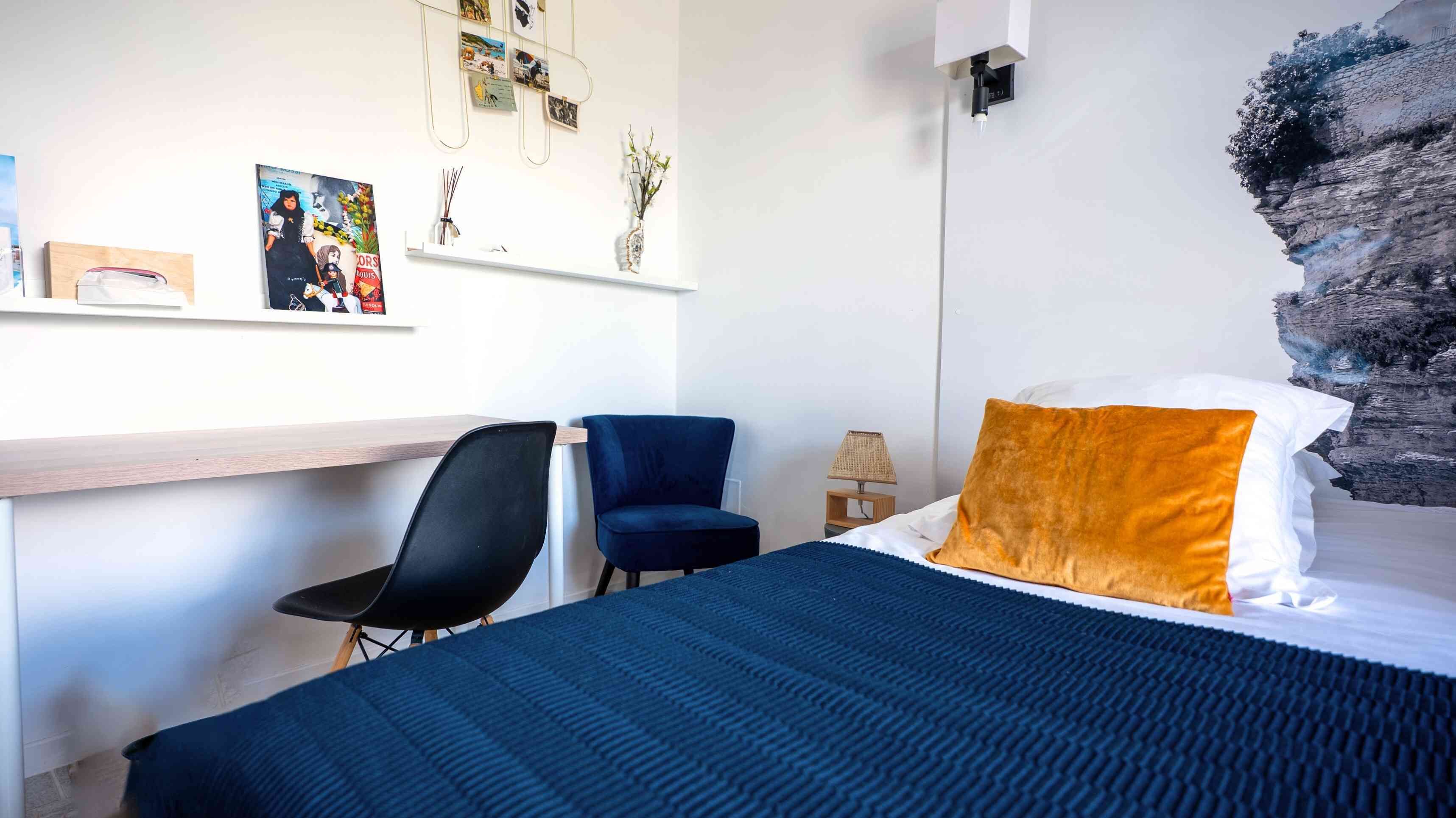 A bedroom with a bed, a desk, two chairs, a lamp, and a wall with photos and a decorative piece.