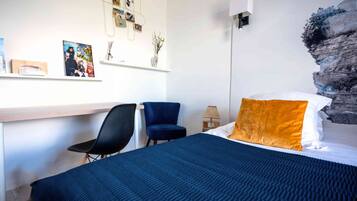 A bedroom with a bed, a desk, two chairs, a lamp, and a wall with photos and a decorative piece.
