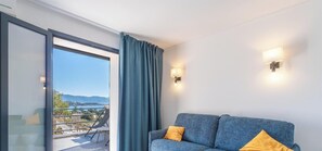 A room with a sofa, a balcony with a view of the sea and mountains, and two wall-mounted lights. - Casa Nova Maison d'hotes (Coggia)
