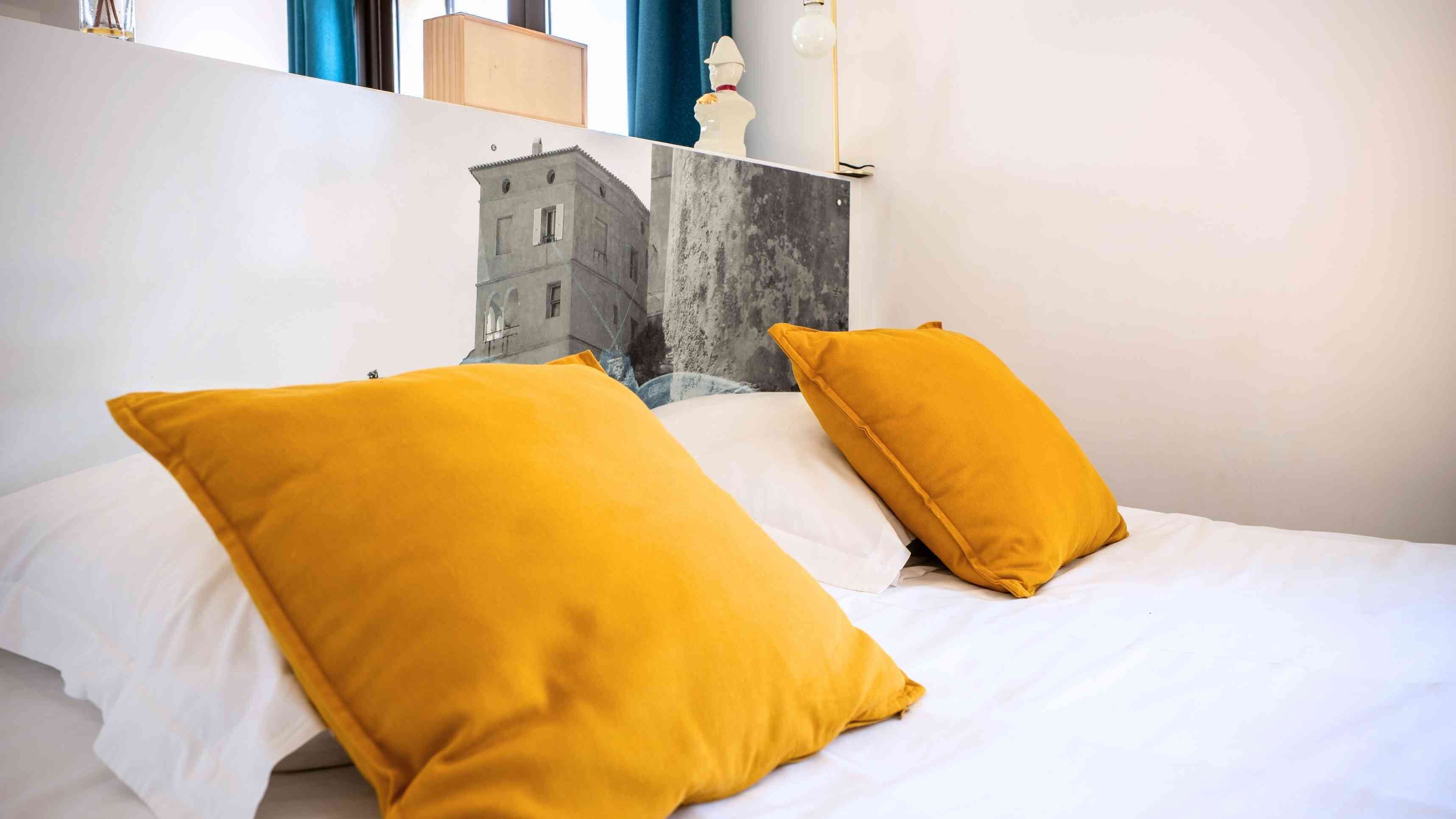 Una cama con ropa blanca y dos almohadas amarillas mostaza, un aplique de pared y un cuadro de un edificio.