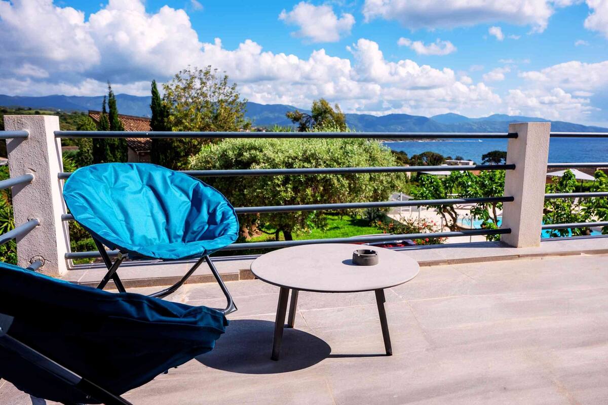 A balcony with a blue chair and a small table, overlooking a pool and mountains.