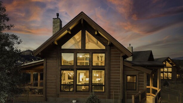 A two-story log cabin with large windows, a stone chimney, and a covered porch. - Chic Retreat – Indoor/Outdoor Fireplace, Private Jacuzzi, Great Location! (Estes Park)