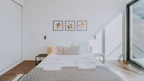 Een moderne hotelkamer met een groot bed, een nachtkastje, en een raam met uitzicht op de stad. Boven het bed hangen drie kunstwerken in lijsten.
