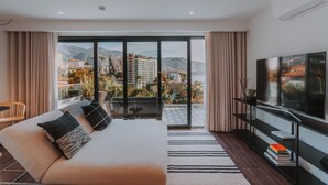 A modern hotel room with a large bed, a TV, a balcony with a view of the city, and a dining area. - FLH Casa Branca Madeira 301 (Funchal)