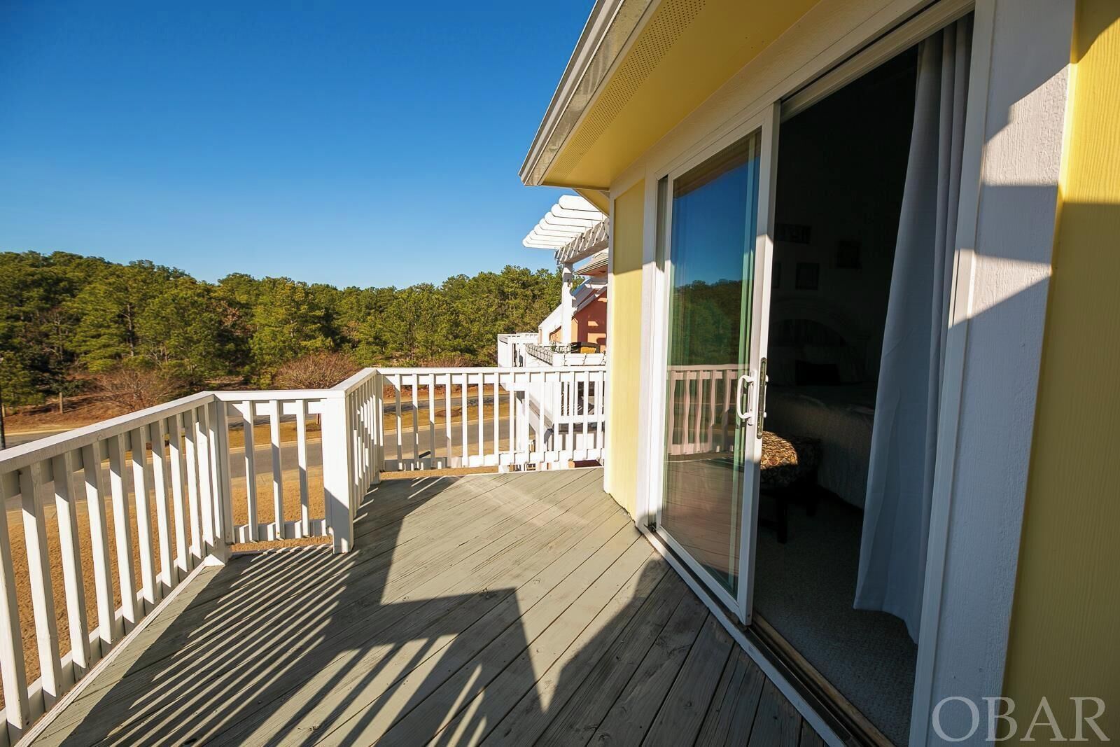 Une terrasse en bois avec une balustrade blanche, une porte vitrée donnant sur une pièce et, au loin, une vue sur les arbres.