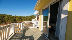 Une terrasse en bois avec une balustrade blanche, une porte vitrée donnant sur une pièce et, au loin, une vue sur les arbres.