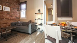 A modern living room with a brick wall, a grey sofa, a small table, and a dining area with a glass table and chairs. - The Sants's balcony (Barcelona)