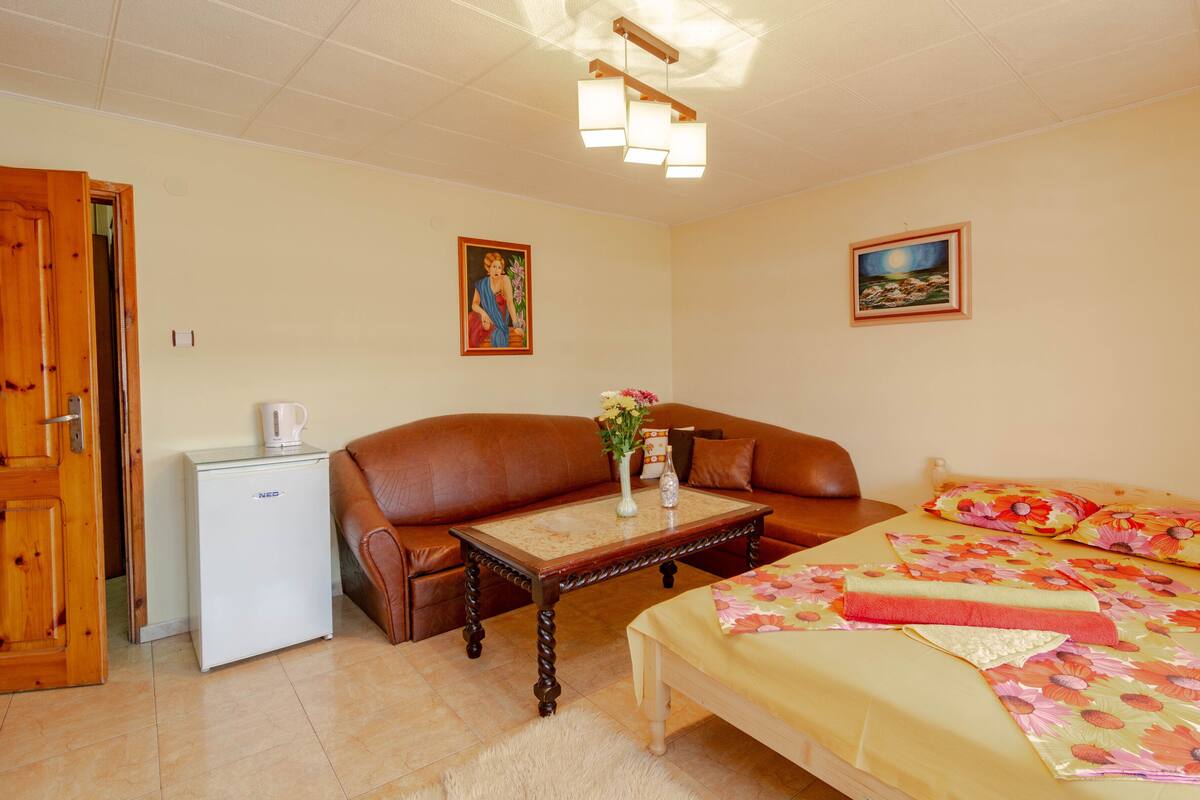 A cozy room with a brown leather sofa, a small white fridge, a wooden table with a vase of flowers, a bed with floral bedding, and two paintings on the walls.