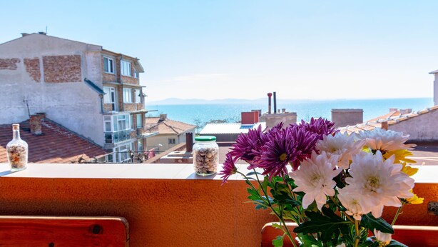 A balcony with flowers, a jar of seashells, and a view of the sea and buildings. - Guest House Fener (Pomorie)