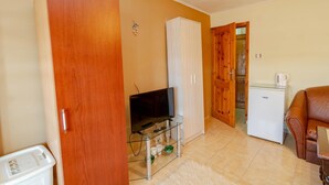 A compact living space with a wooden door, a television on a glass stand, a white cabinet, a refrigerator, and a brown leather sofa. - Guest House Fener (Pomorie)
